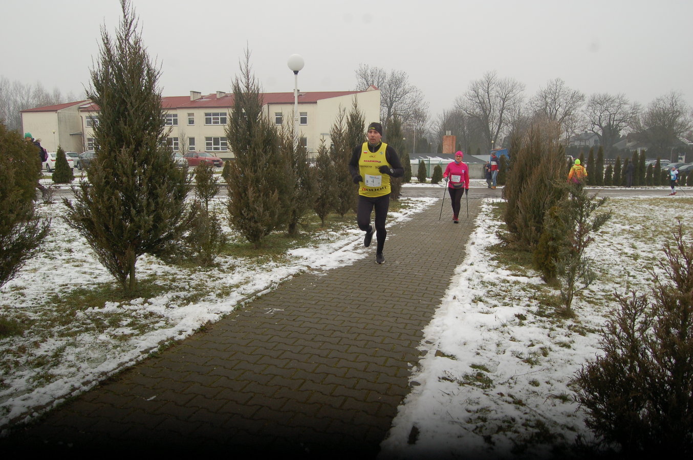 zawodnicy na trasie biegu i marszu , trawa jest pokryta cienka warstwą śniegu, chodnik jest czarny
