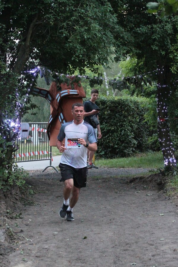 zawodnik biegnie ścieżką po parku, w tle maskotka wiatraczka i młody mężczyzna